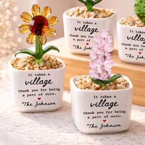 May include: Four decorative glass plants in white square ceramic pots. Each pot features the black text: It takes a village, thank you for being a part of ours, and The Johnson. The plants include a yellow flower, white flowers, a green cactus, and pink beads.
