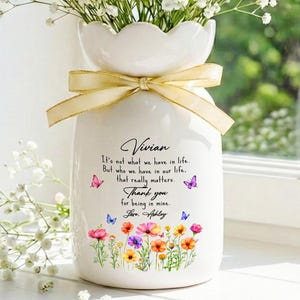 May include: A white ceramic vase with a scalloped rim and a beige ribbon tied around the neck. The vase features a printed floral design, colourful butterflies, and the text: Vivian, It's not what we have in life. But who we have in our life. That really matters. Thank you for being in mine. Love, Ashley.
