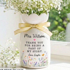 May include: A white ceramic vase with a scalloped rim, filled with white baby's breath flowers. A gold ribbon is tied around the neck. The vase features the text Mrs. Wilson, Thank you for being a part of my story, Love, Layla, and a small pink heart.