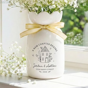 May include: A white ceramic vase with a scalloped rim, tied with a cream-colored ribbon. The front features a black graphic of a house with the text A NEW CHAPTER BEGINS, Jordan & Ashton, 1234 HAPPY STREET, and 2026, filled with white baby's breath.