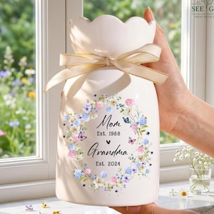 May include: A cream-colored ceramic vase with a scalloped rim and a beige ribbon tied in a bow around the neck. The front features a floral wreath graphic with butterflies and the text: Mom Est. 1988, a heart symbol, Grandma Est. 2024.