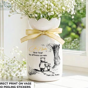 May include: White ceramic vase with a scalloped top, filled with white flowers. A gold ribbon is tied around the neck. The vase features a black and white illustration of a bear and a piglet with the text "Never forget the difference you make."