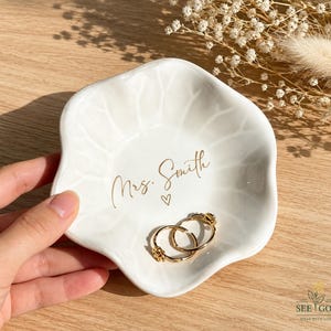 May include: A white, scalloped-edge ceramic trinket dish featuring a subtle leaf-vein texture. The center is inscribed with the gold text "Mrs. Smith" above a small heart symbol. Two gold rings are resting inside the dish on the smooth surface.