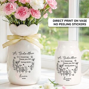 May include: A white ceramic vase with a scalloped rim and a gold ribbon, filled with pink and white carnations, alongside a matching white ceramic mug. Both items feature black floral illustrations and the text: A Godmother is a blessing. Thank you for being mine. Laura 2026.