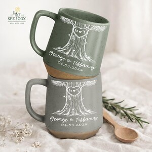May include: Two stacked ceramic mugs with a tree design and the names "George & Tiffany" and the date "04.03.2026" in white. The mugs are gray-green with a brown base and a large handle. The tree design includes a heart with the initials "G T".