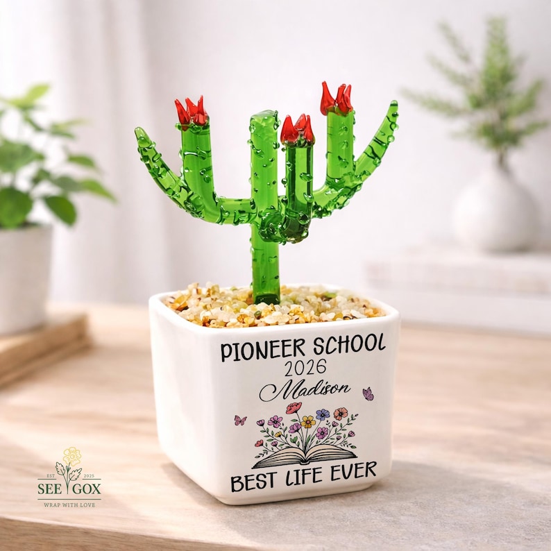 May include: A green glass cactus with red flowers sits in a white square ceramic pot filled with small stones. The pot features the text PIONEER SCHOOL 2026, the name Madison, a floral illustration above an open book, and BEST LIFE EVER.