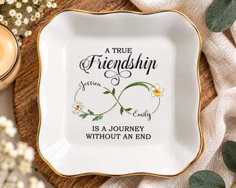 May include: A square white ceramic trinket dish with a gold-painted scalloped rim. The center features an infinity symbol graphic with floral accents, the names Jessica and Emily, and the text A TRUE Friendship IS A JOURNEY WITHOUT AN END.