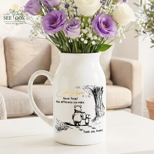 May include: White ceramic vase with a handle, filled with purple and white flowers. The vase features a black and gold illustration of a bear and a smaller animal, with the text "Never forget the difference you make". The vase has the text "Thank you, Avery".