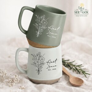 May include: Two ceramic mugs, one sage green and one white, stacked. Each mug features a floral illustration and the text "Aunt Laura Est. 2026." The mugs have a speckled finish and a natural-colored base.