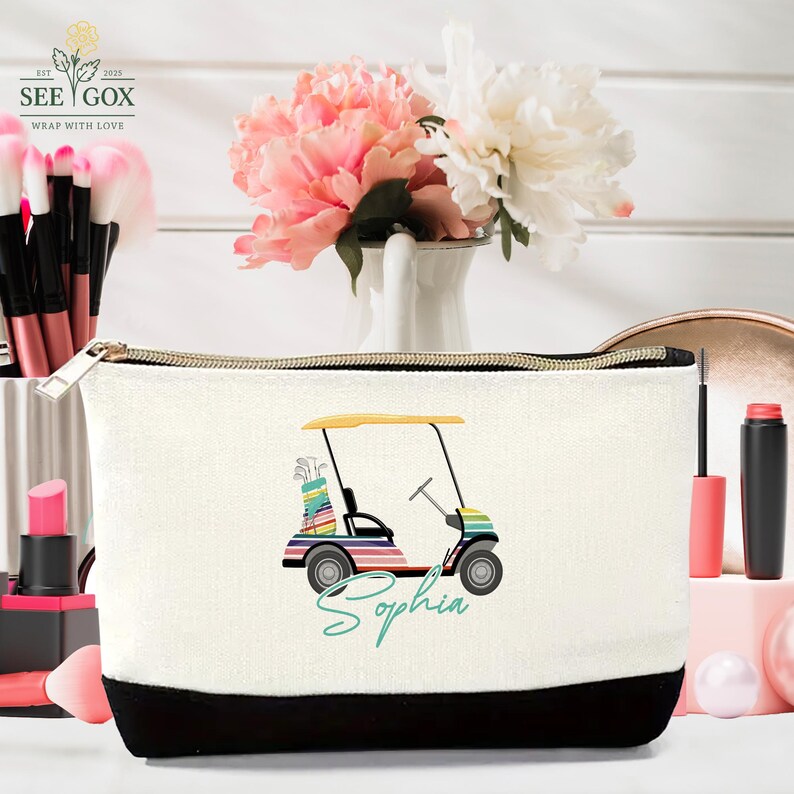 Puede incluir: Bolsa de cosm&eacute;ticos de lona blanca con base negra y cremallera dorada. La bolsa presenta un dise&ntilde;o de carrito de golf colorido con el nombre "Sophia" en escritura turquesa. Decorada con brochas de maquillaje y flores.