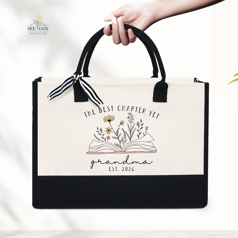 May include: A tote bag with black handles and a black bottom panel. The bag features the text "THE BEST CHAPTER YET grandma EST. 2026" above an illustration of an open book with flowers.