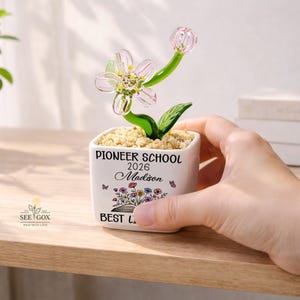 May include: A white square ceramic planter filled with small tan pebbles and a glass flower sculpture. The planter features the text PIONEER SCHOOL 2026, the name Madison, and a colorful floral graphic with butterflies printed on the front surface.