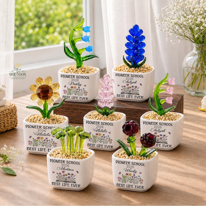 May include: Seven small white ceramic square planters, each containing a glass flower sculpture and light-colored pebbles. Each planter features the text PIONEER SCHOOL 2026, a unique name, and a floral graphic above the text BEST LIFE EVER.