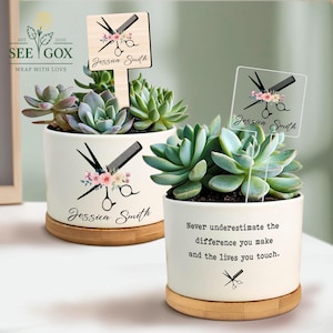 May include: Two white ceramic succulent planters with wooden bases. Each planter features a floral design with scissors and a comb, and the name "Jessica Smith." One planter has a wooden sign with the same design. The other planter has the text "Never underestimate the difference you make and the lives you touch."