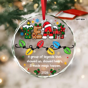 May include: A round clear glass Christmas ornament with a red ribbon hanger. The design features the word TEAM in festive patterns, a string of colorful light bulbs with names Emily, Robert, James, Kevin, William, and Mary, and the text A group of legends that showed up, showed heart, & made magic happen. 2026 is printed at the bottom.