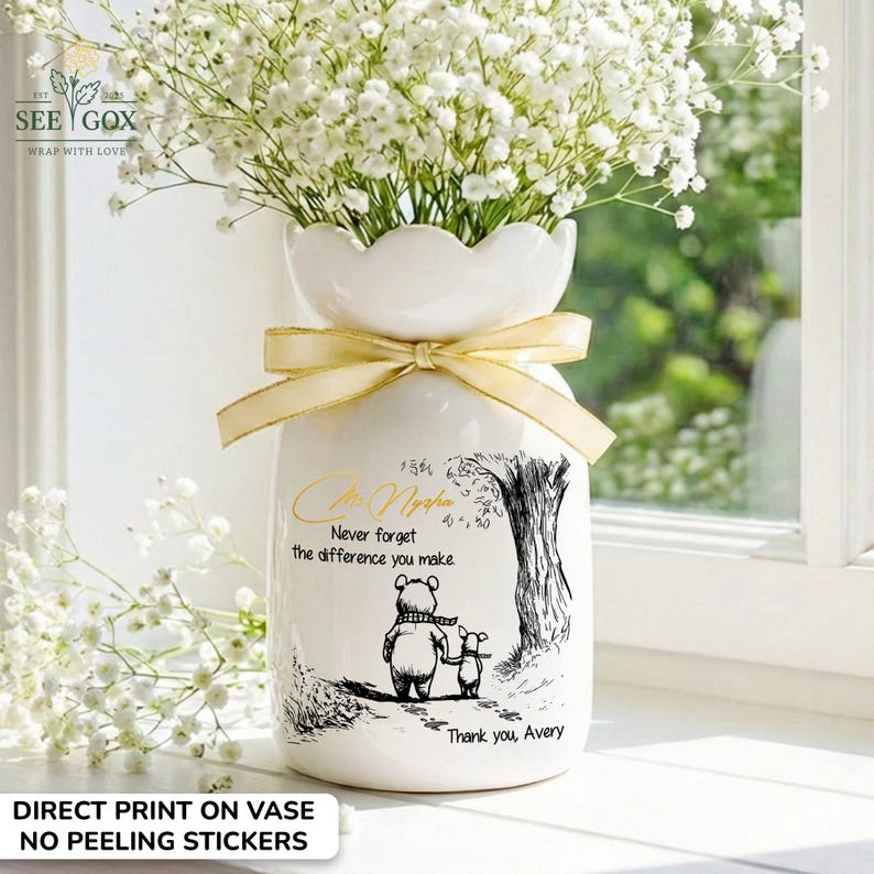 May include: White ceramic vase with a scalloped top, filled with white flowers. A gold ribbon is tied around the neck. The vase features a black and white illustration of a bear and a piglet with the text "Never forget the difference you make."