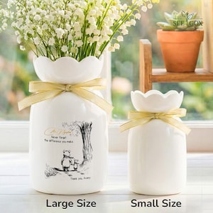 May include: Two white ceramic vases with scalloped rims and gold ribbon accents. The larger vase features a Winnie the Pooh illustration with text that reads "Never forget the difference you make. Thank you, Avery." The vases are labeled "Large Size" and "Small Size".