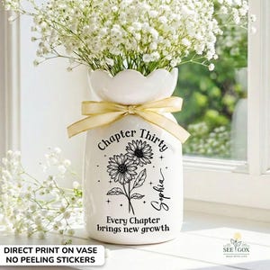 May include: A white ceramic vase with a floral design and the text "Chapter Thirty Sophia". The vase is adorned with a gold ribbon and filled with white flowers. The text "Every Chapter brings new growth" is also printed on the vase.