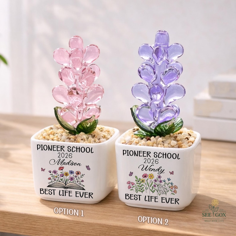May include: Two square white ceramic planters filled with small pebbles. One features a pink glass heart-shaped flower, the other a purple one. Both are printed with 'PIONEER SCHOOL 2026', a name, a floral illustration, and 'BEST LIFE EVER'.