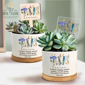 May include: Two white ceramic succulent pots with wooden bases, each featuring a personalized design for "Lauren." The design includes hairstyling tools and the text "a scissor wielding, miracle working, creative genius who touches more hearts than hair."