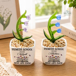 May include: Two white square ceramic planters filled with small pebbles, each holding a glass lily of the valley plant. One has purple flowers, the other blue. Both feature the text PIONEER SCHOOL 2026, a name, and BEST LIFE EVER.