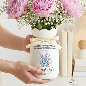 May include: A white ceramic vase with a scalloped rim, tied with a cream ribbon, holding pink peonies and baby's breath. The vase features a blue stethoscope graphic, floral accents, and the text: Never underestimate the difference you have made.