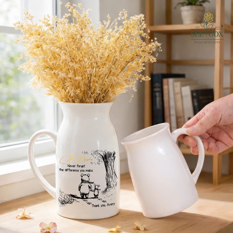 May include: White ceramic pitcher with a handle, holding dried yellow flowers. The pitcher has a black and white illustration of a bear and a mouse with the text "Never forget the difference you make. Thank you, Avery."
