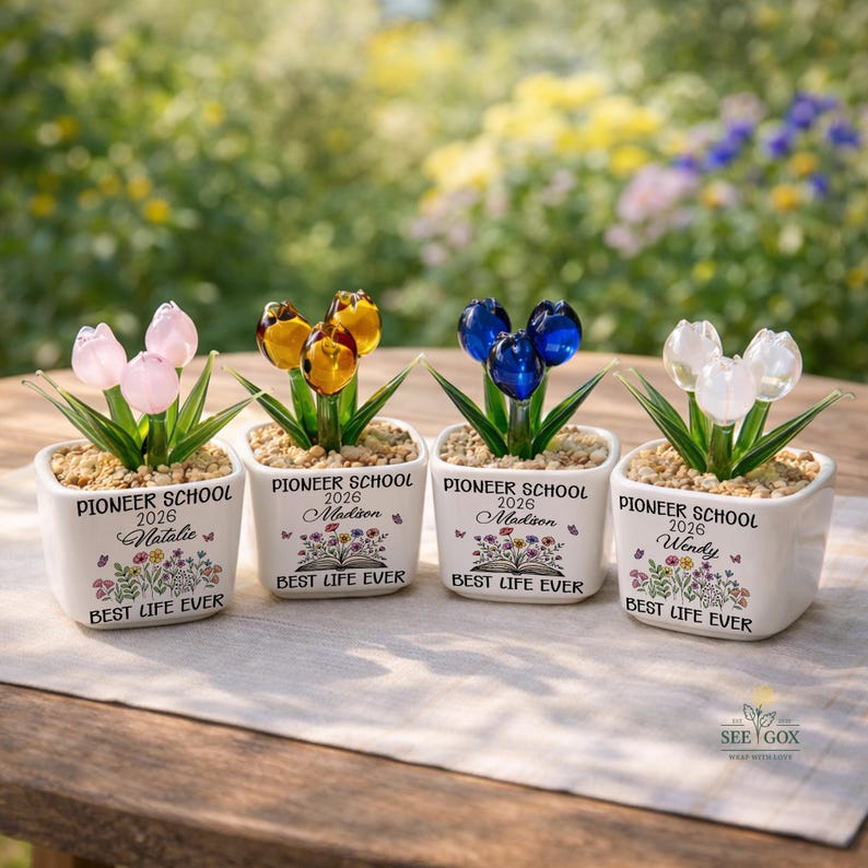 May include: Four square white ceramic planters containing glass tulip flowers in pink, amber, blue, and white. Each planter features the text PIONEER SCHOOL 2026, a name, a floral illustration, and the text BEST LIFE EVER, filled with small pebbles.