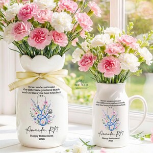 May include: A white ceramic vase and a matching white ceramic mug, both filled with pink and white carnations. Each item features a blue stethoscope graphic, floral accents, and the text: Never underestimate the difference you have made and the lives you have touched. Amanda, RN. Happy Retirement 2026.