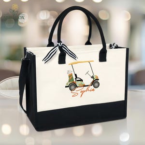 Puede incluir: Bolso tote blanco y negro con asas negras y una correa para el hombro. El bolso presenta un gr&aacute;fico de carrito de golf con el nombre "Sophia" en cursiva. El bolso tiene una base negra y un detalle de cinta a rayas.