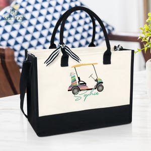 Puede incluir: Un bolso de mano de lona con asas negras y una base negra. El bolso presenta un gr&aacute;fico de carrito de golf colorido y el nombre "Sophia" en escritura verde. Una cinta a rayas blancas y negras est&aacute; atada a una de las asas.