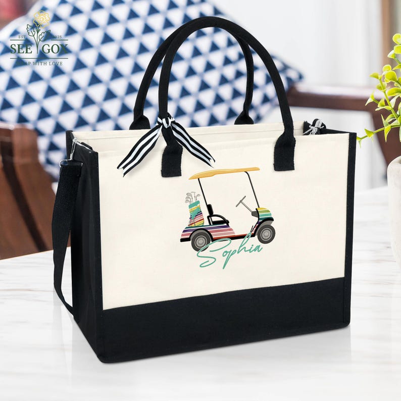 Puede incluir: Un bolso de mano de lona con asas negras y una base negra. El bolso presenta un gr&aacute;fico de carrito de golf colorido y el nombre "Sophia" en escritura verde. Una cinta a rayas blancas y negras est&aacute; atada a una de las asas.