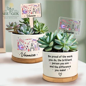 May include: Two white ceramic succulent pots with wooden bases. Each pot features a nail salon theme with tools and the name "Veronica". One pot has a wooden sign, the other a clear sign. The pots contain green succulents.