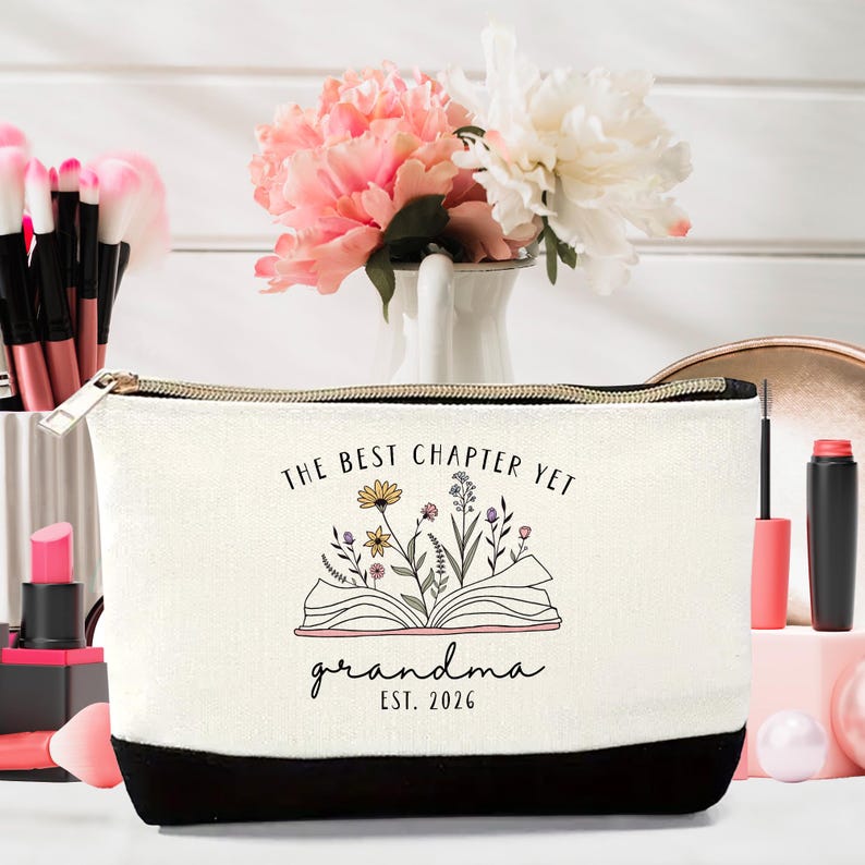 May include: A white canvas cosmetic bag with a black base and a gold zipper. The bag features the text "THE BEST CHAPTER YET grandma EST. 2026" with a floral book design. Makeup brushes and flowers are in the background.