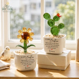 May include: Two glass plant figurines in white square ceramic pots. One features a yellow crystal sunflower, the other a green crystal cactus with a red flower. Both pots are printed with the text A new Chapter, a quote, and the name Emma.