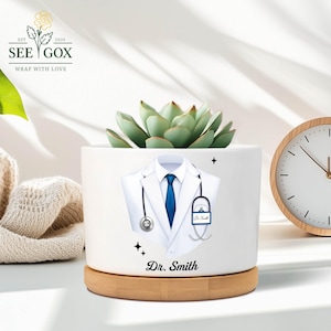 May include: White ceramic planter featuring a doctor's coat design, a blue tie, and a stethoscope. The planter holds a succulent plant and rests on a wooden base. The name "Dr. Smith" is printed on the planter.