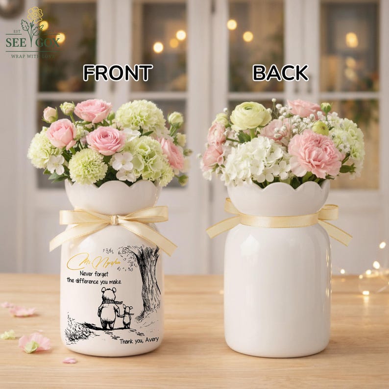 May include: Two white ceramic vases, each holding a floral arrangement of pink and white flowers. The front vase has a Winnie the Pooh illustration and text that reads "Never forget the difference you make. Thank you, Avery."