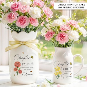 May include: Two white ceramic vases filled with pink and white carnations. The larger vase has a yellow ribbon and text reading Chapter FORTY every chapter brings new growth Anna. The smaller mug-style vase features text reading Chapter FORTY every chapter brings new growth Jessica with yellow flowers.
