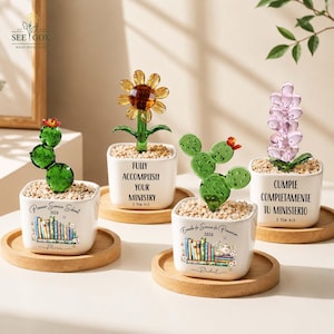 May include: Four square white ceramic pots on wooden coasters, each containing a glass plant sculpture. Two pots feature illustrations of books and text, while two feature text only. The glass plants include a green cactus, a sunflower, and a lavender flower.