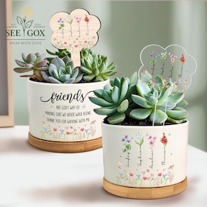 May include: Two white ceramic succulent planters with wooden bases. One planter has the word "friends" and a quote. The other has names. Both planters have a floral design and a wooden flower-shaped stake.