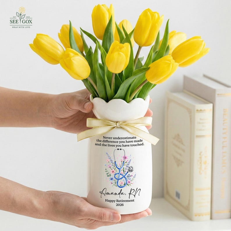 May include: A white ceramic vase with a scalloped rim and a beige ribbon, filled with yellow tulips. The vase features a blue stethoscope graphic, floral accents, and the text: Never underestimate the difference you have made and the lives you have touched. Amanda, RN. Happy Retirement 2026.