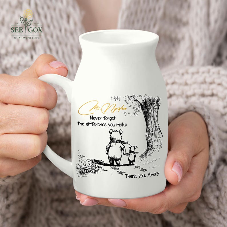 May include: White ceramic mug with a handle, featuring a black and gold illustration of a bear and a piglet walking together. The mug has the text "Mrs. Nypha, Never forget the difference you make. Thank you, Avery."