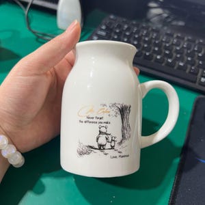 May include: White ceramic mug with a handle, featuring a Winnie the Pooh illustration and the text "Never forget the difference you make." The mug has a curved shape and a wide opening, perfect for coffee or tea.