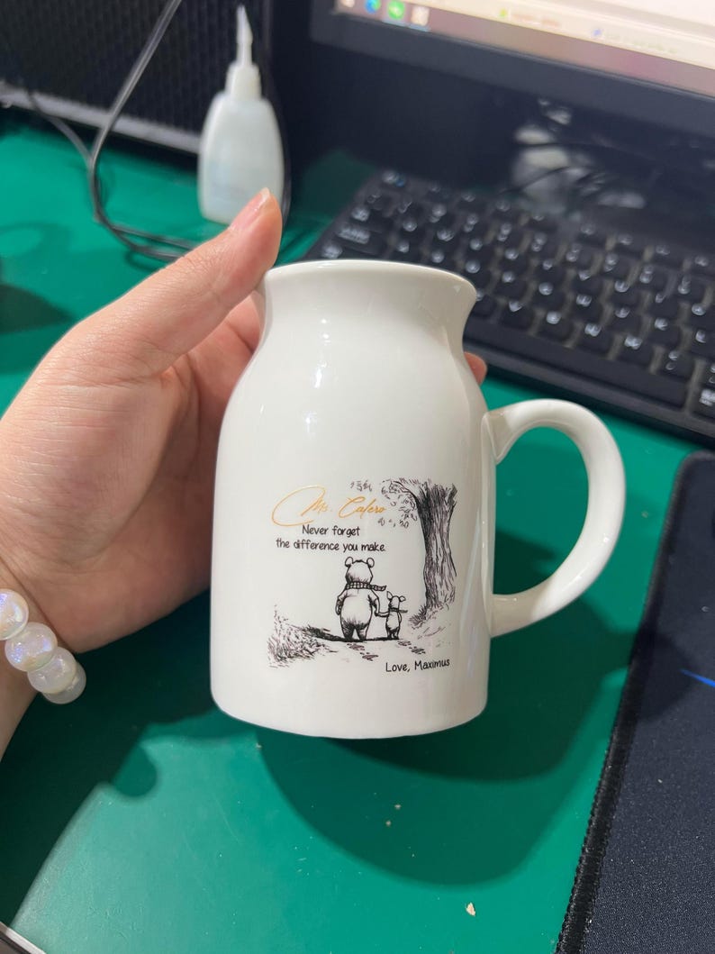 May include: White ceramic mug with a handle, featuring a Winnie the Pooh illustration and the text "Never forget the difference you make." The mug has a curved shape and a wide opening, perfect for coffee or tea.