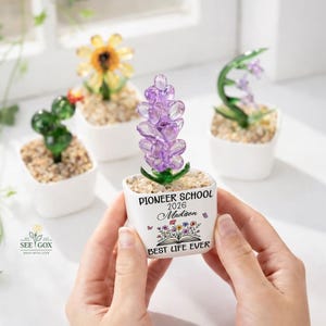 May include: A small white square planter containing a purple glass flower, with text reading PIONEER SCHOOL 2026 Madison BEST LIFE EVER and a floral graphic. Other glass plants in white pots are visible in the blurred background.