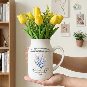 May include: A white ceramic pitcher vase filled with yellow tulips and green foliage. The front features a blue stethoscope graphic surrounded by flowers, with the text: Never underestimate the difference you have made and the lives you have touched. Amanda, RN. Happy Retirement 2026.