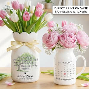 May include: Two white ceramic vases with floral arrangements. One vase holds pink and white tulips, the other holds pink peonies. The vases have text and a gold ribbon. One vase has a calendar.