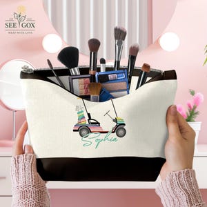 Puede incluir: Un neceser de lona con base y cremallera negras, lleno de brochas y productos de maquillaje. La bolsa presenta un dise&ntilde;o de carrito de golf colorido con el nombre "Sophia" en una fuente cursiva.