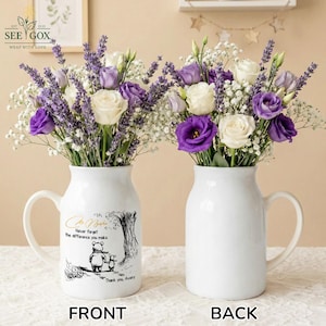 May include: Two white ceramic vases with handles, each filled with a bouquet of white, purple, and lavender flowers. One vase has a Winnie the Pooh illustration and text that reads "Never forget the difference you make."