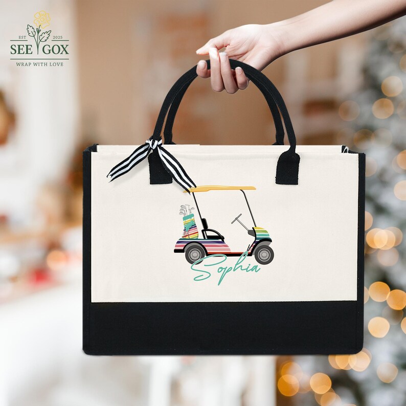 Puede incluir: Un bolso de mano de lona con asas negras y un panel inferior negro. El bolso presenta un gr&aacute;fico de carrito de golf colorido con el nombre "Sophia" en escritura turquesa. El bolso es sostenido por una persona.